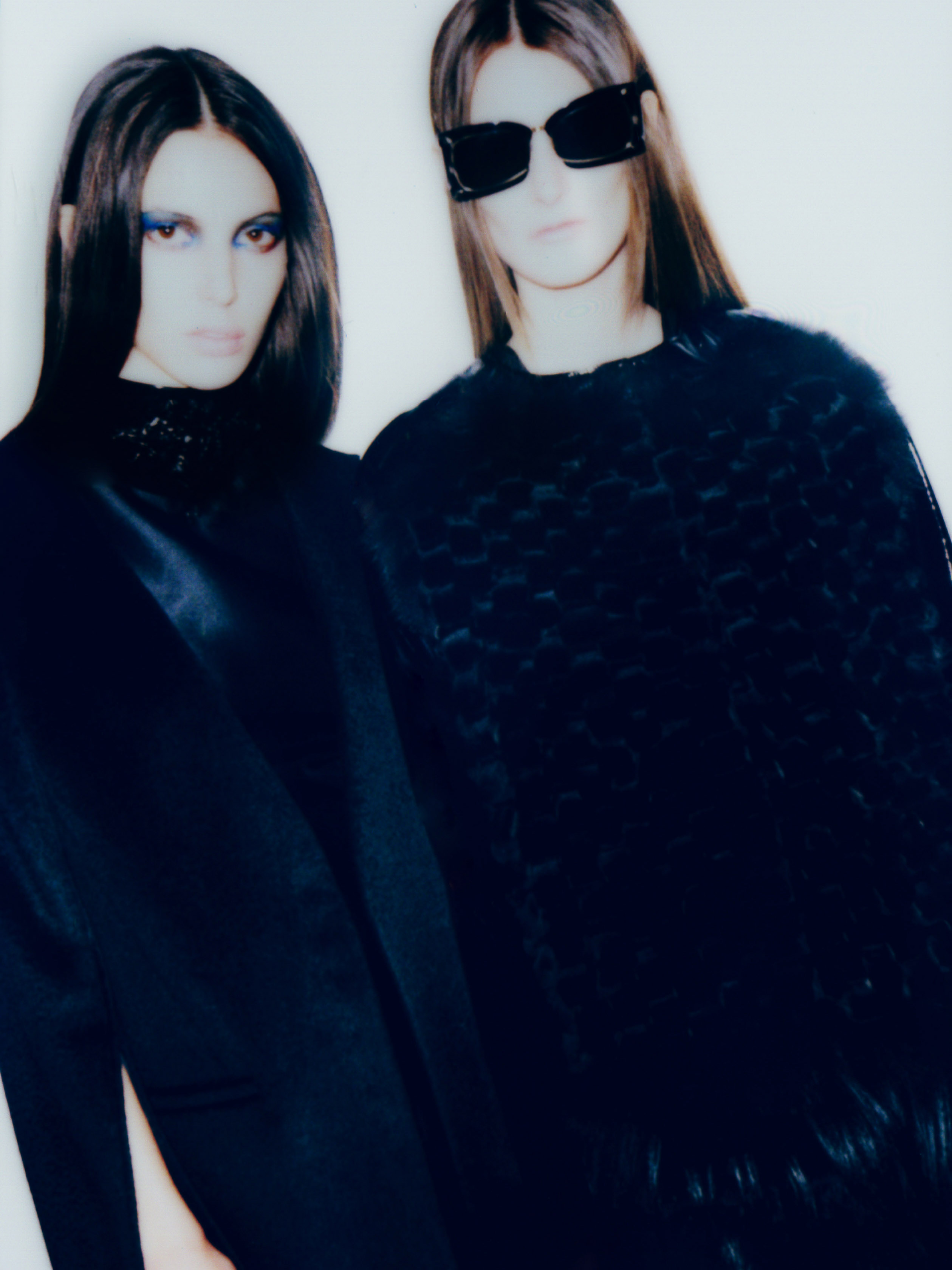 Two women pose indoors against a white backdrop, showcasing black high-fashion outfits and statement sunglasses in a studio fashion shoot.