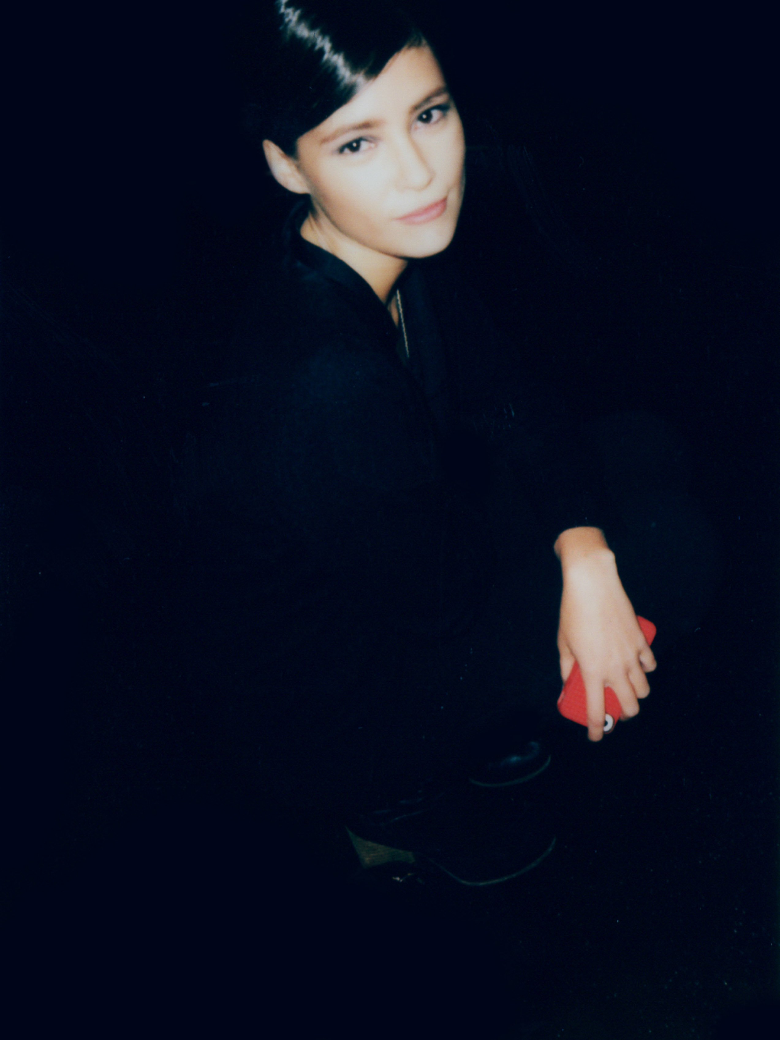 Portrait of a woman with dark hair in dark clothing, crouching and holding a red cell phone, set against a dark, shadowy background.