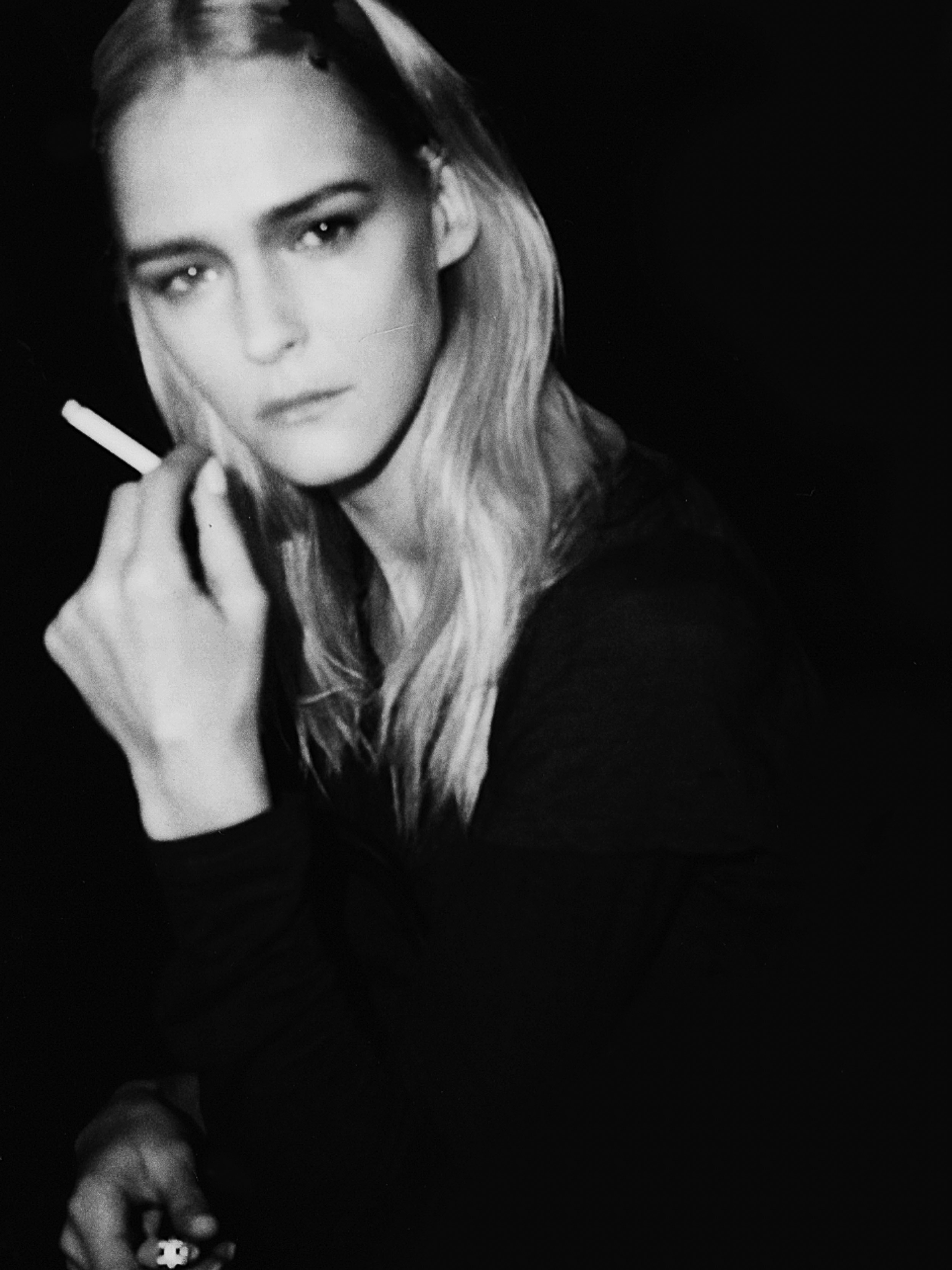 Monochrome portrait of a thoughtful adult woman with long blond hair holding a lit cigarette in a dark, contrasted indoor setting.