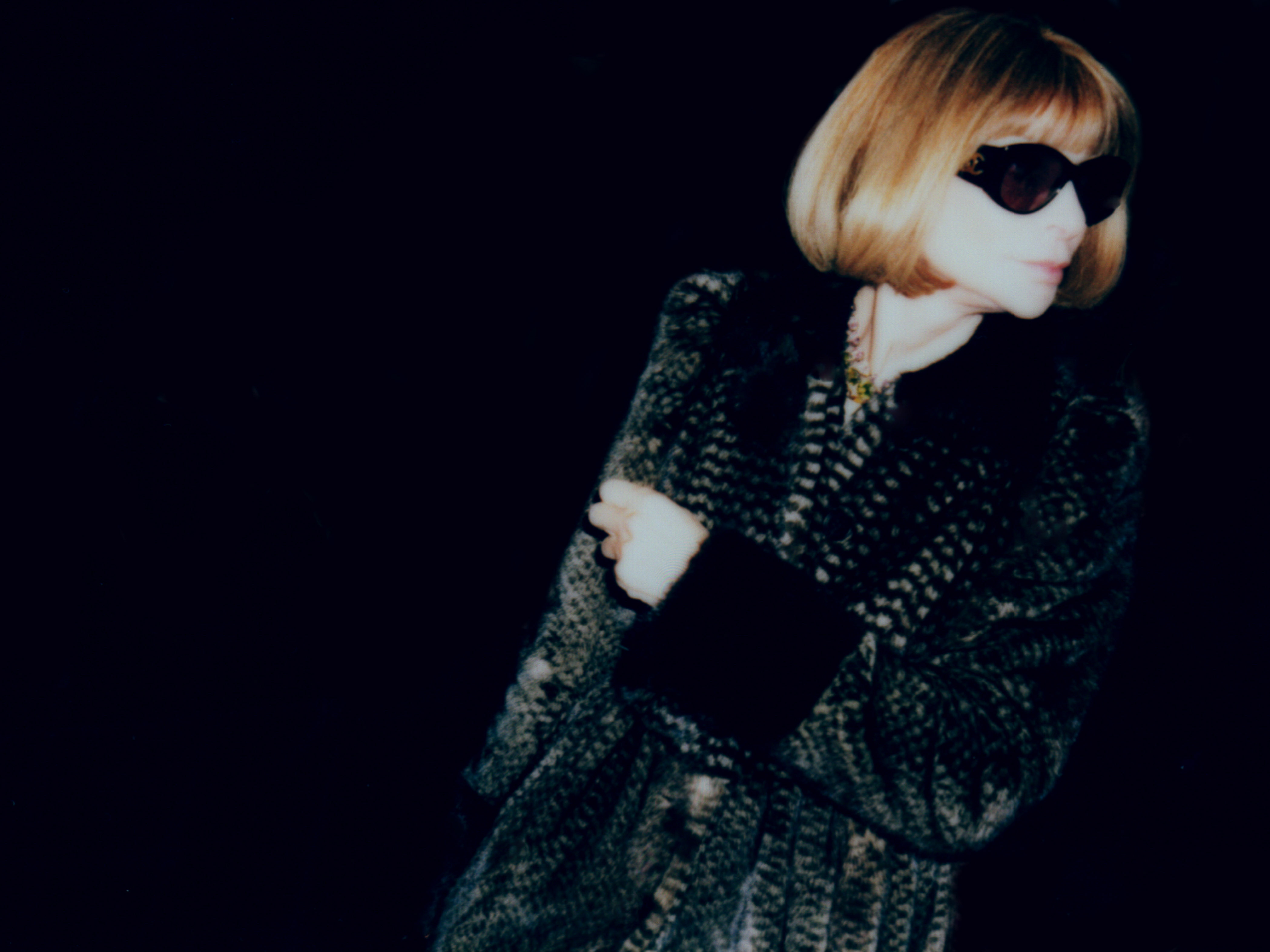 A stylish woman with blonde bob, wearing sunglasses and patterned coat, poses against a dark backdrop in a fashion editorial.