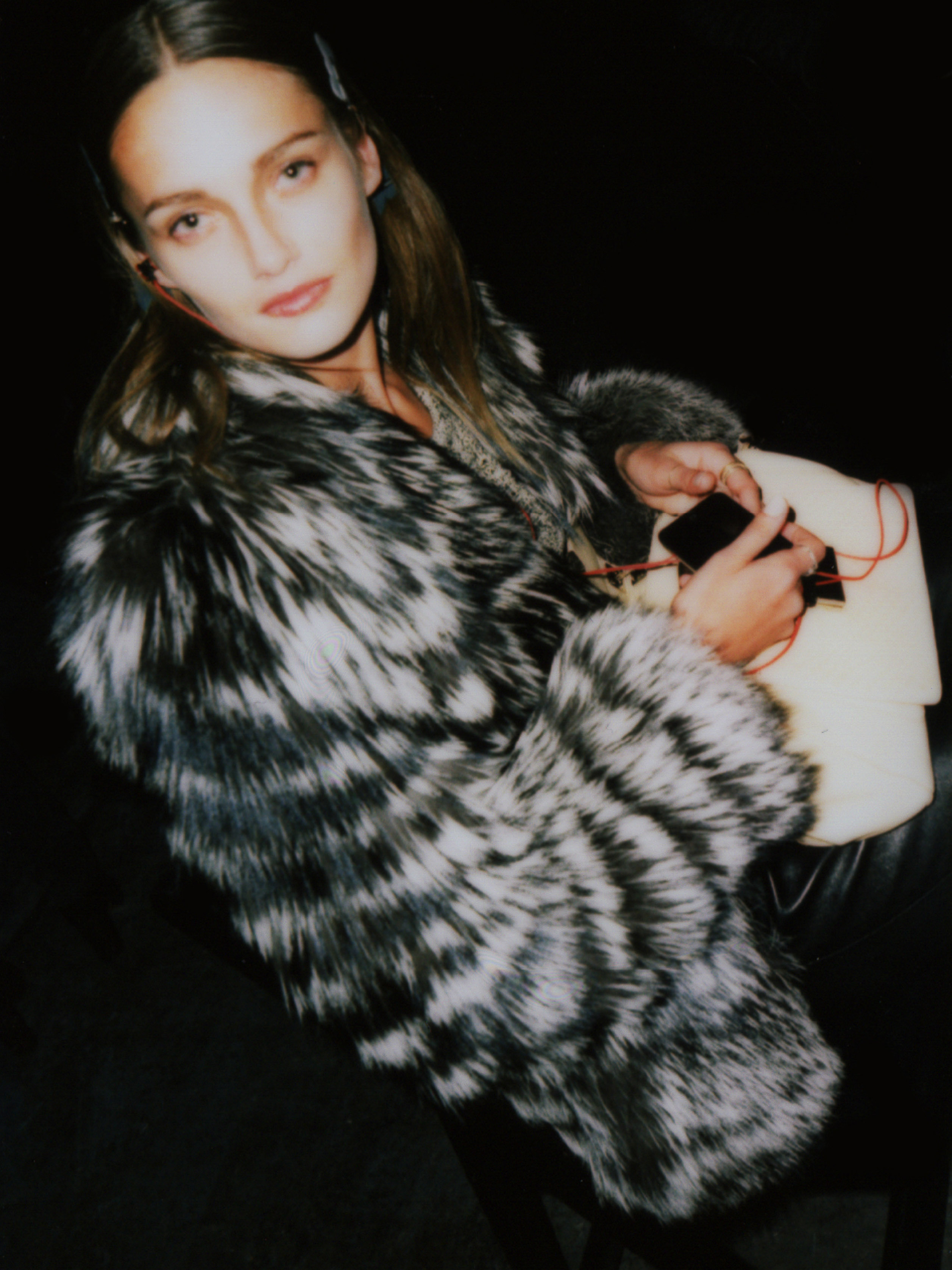 Portrait of a young adult female with fair skin and light brown hair wearing a black and white fur coat, holding a black cellphone.