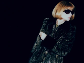 Anna Wintour wearing sunglasses and a patterned coat against a dark background, photographed by Cameron Krone for New York Times Style.