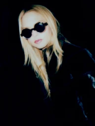 Portrait of a blonde woman wearing dark round sunglasses and a black jacket against a dark background