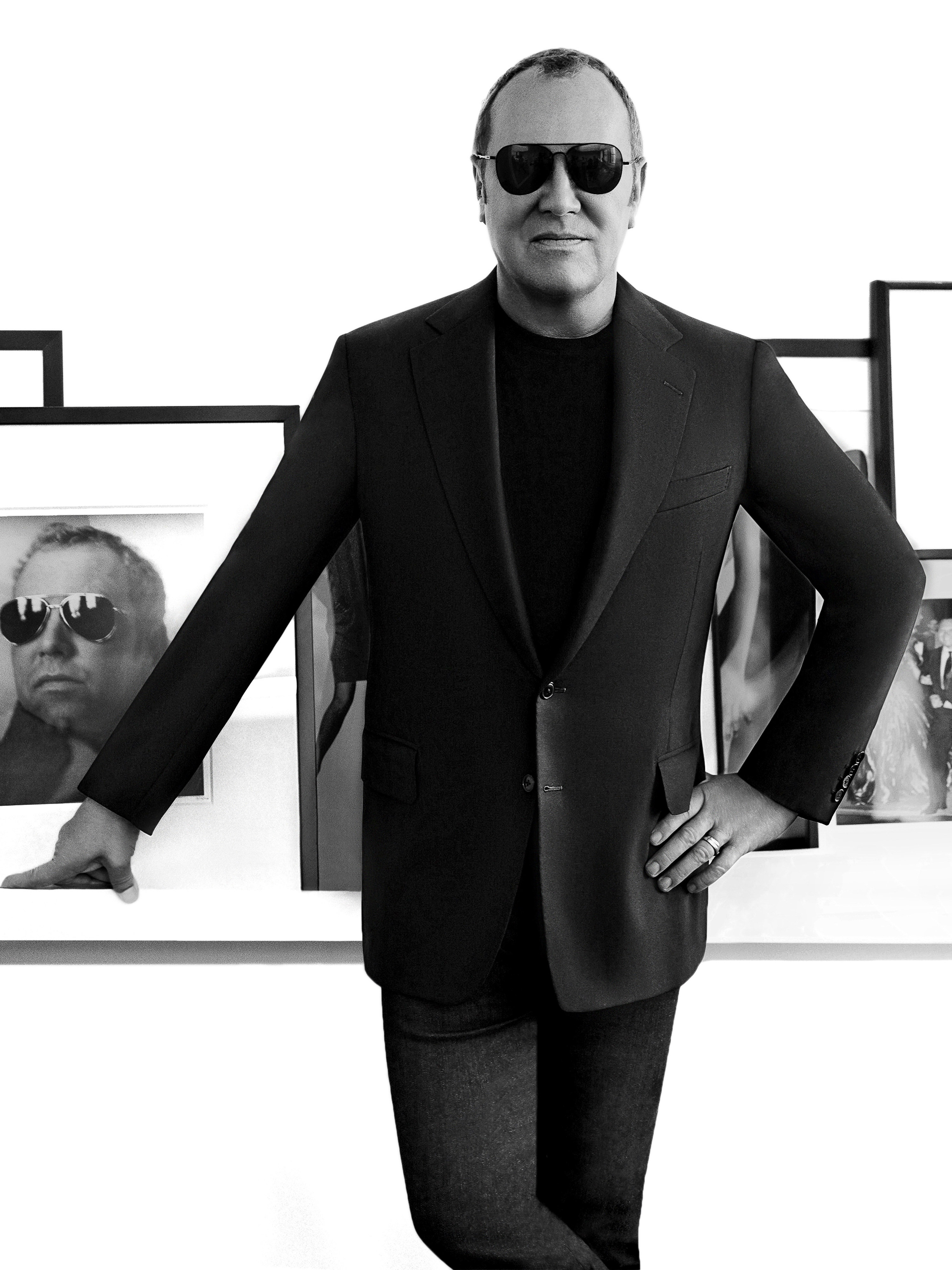 Black and white portrait of an adult male wearing sunglasses, a blazer, and jeans in front of framed photos on a white background.