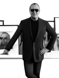 Monochrome portrait of Michael Kors in a dark blazer and sunglasses standing before framed artwork