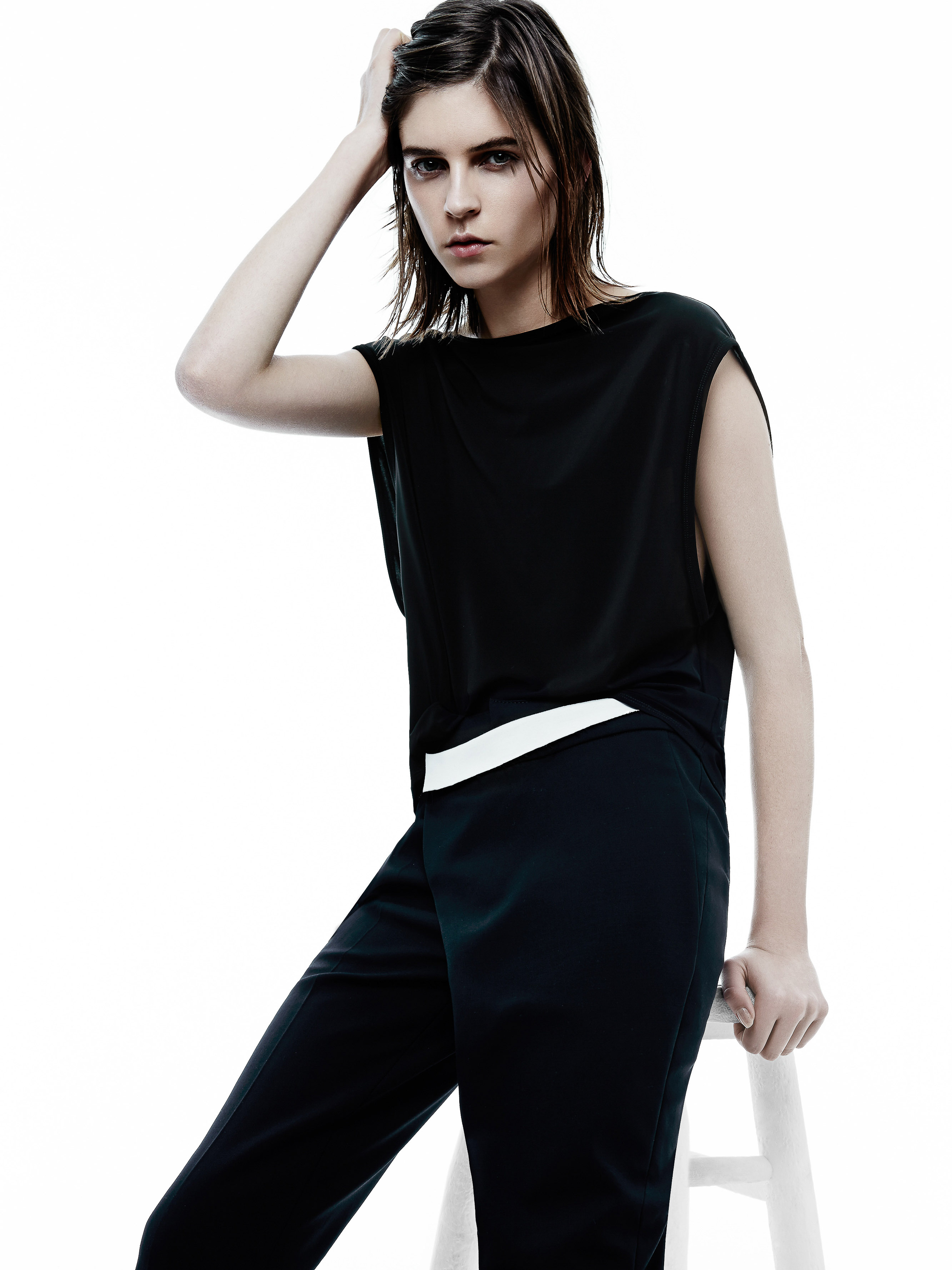 Fashion shot of an adult woman with dark hair in a sleeveless top and trousers sitting on a white stool in a bright photo studio.