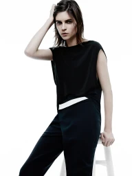 Model seated on a white stool wearing a black sleeveless top and black trousers with a white waistband against a white background.