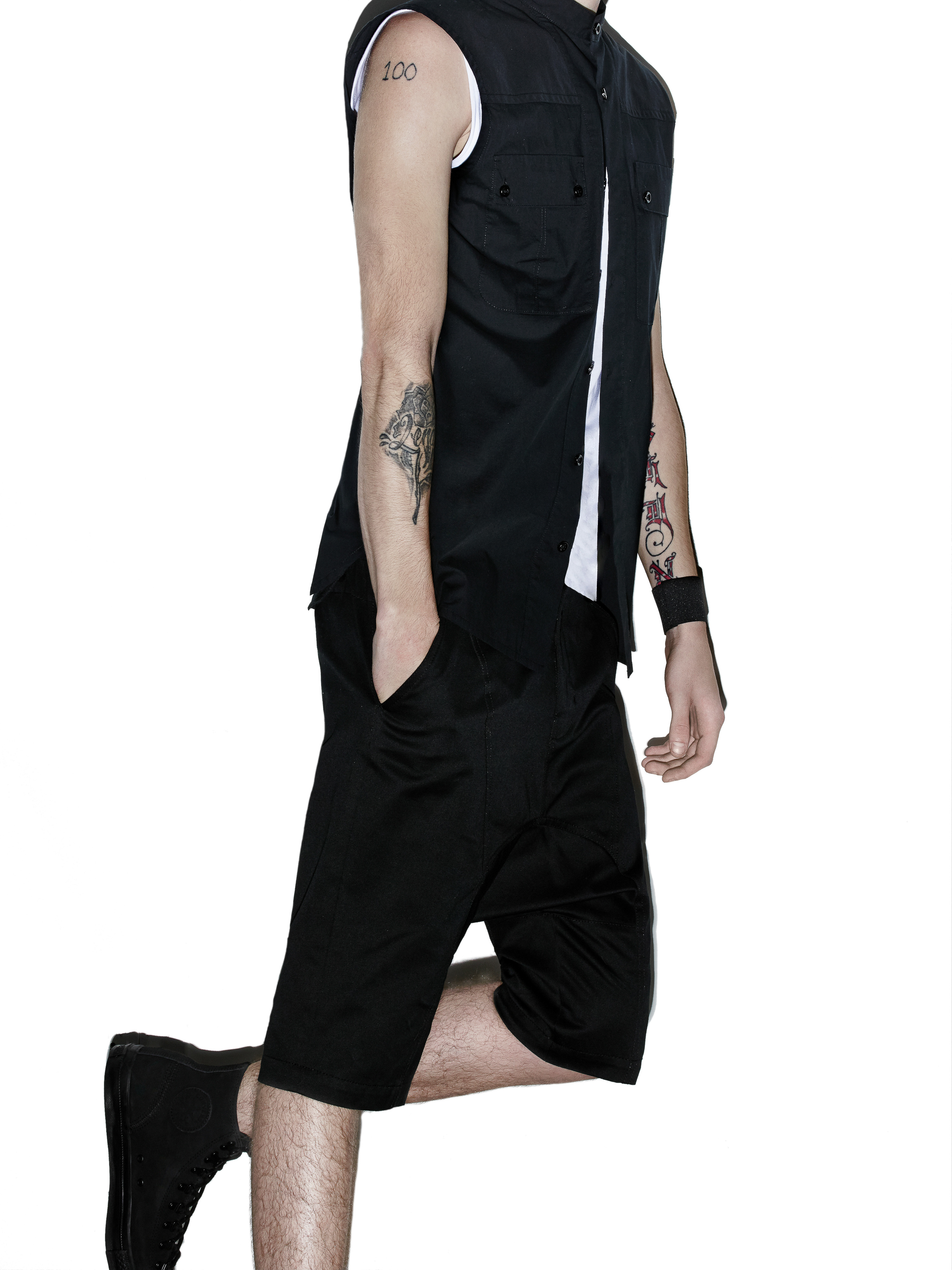 Model wearing a sleeveless black collared shirt, black knee-length shorts, and black sneakers against a white seamless background.