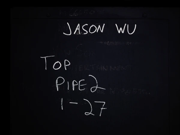 Blackboard with white handwritten text featuring 'Jason Wu,' 'Top,' 'Pipe 2,' and '1-27,' offering a stark, monochromatic visual.