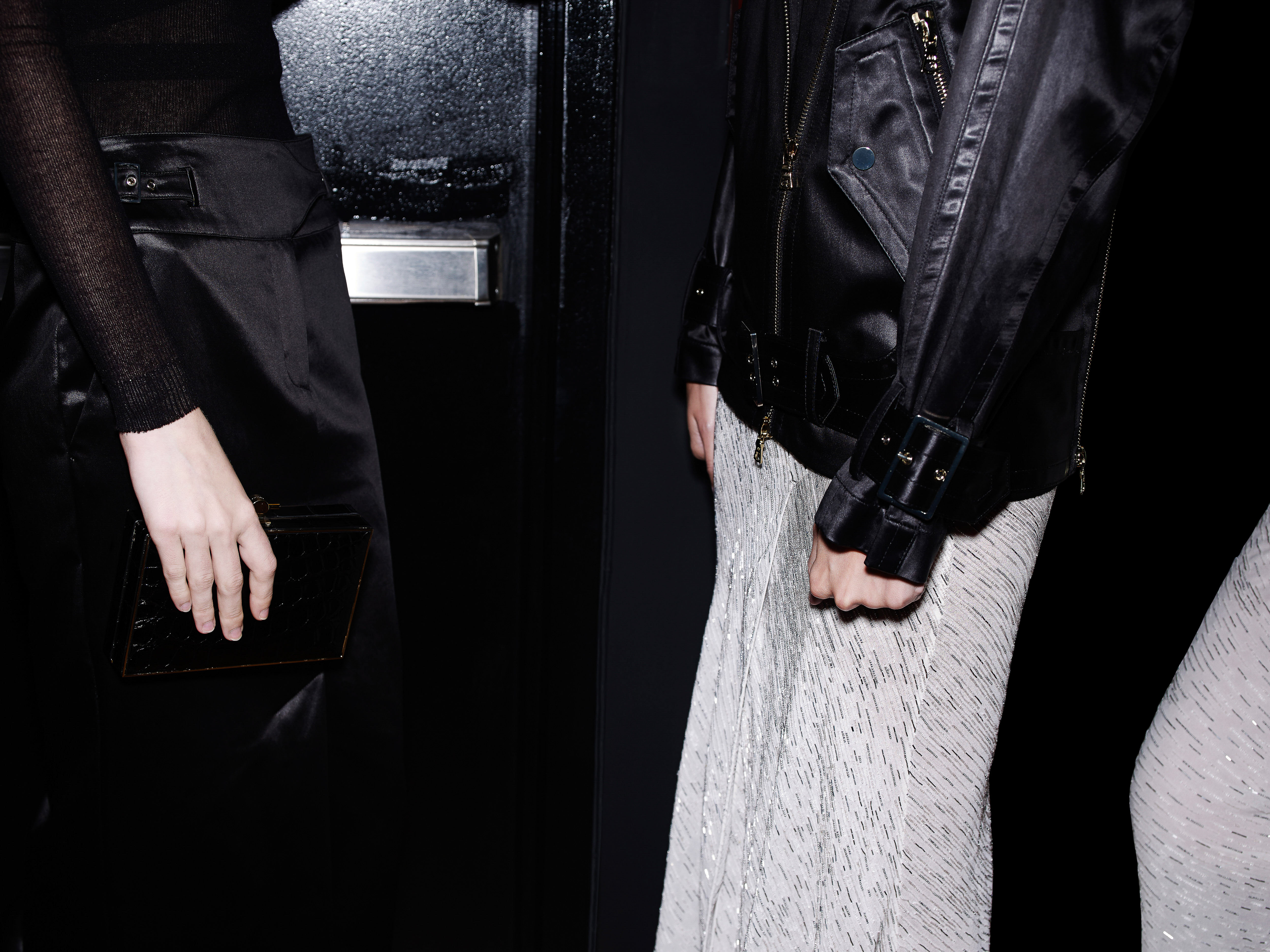 Two women pose indoors, showcasing black shiny jackets and high-waisted skirts against a dark backdrop with a partially visible metal door.