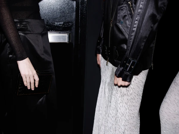 Two women pose indoors, showcasing black shiny jackets and high-waisted skirts against a dark backdrop with a partially visible metal door.