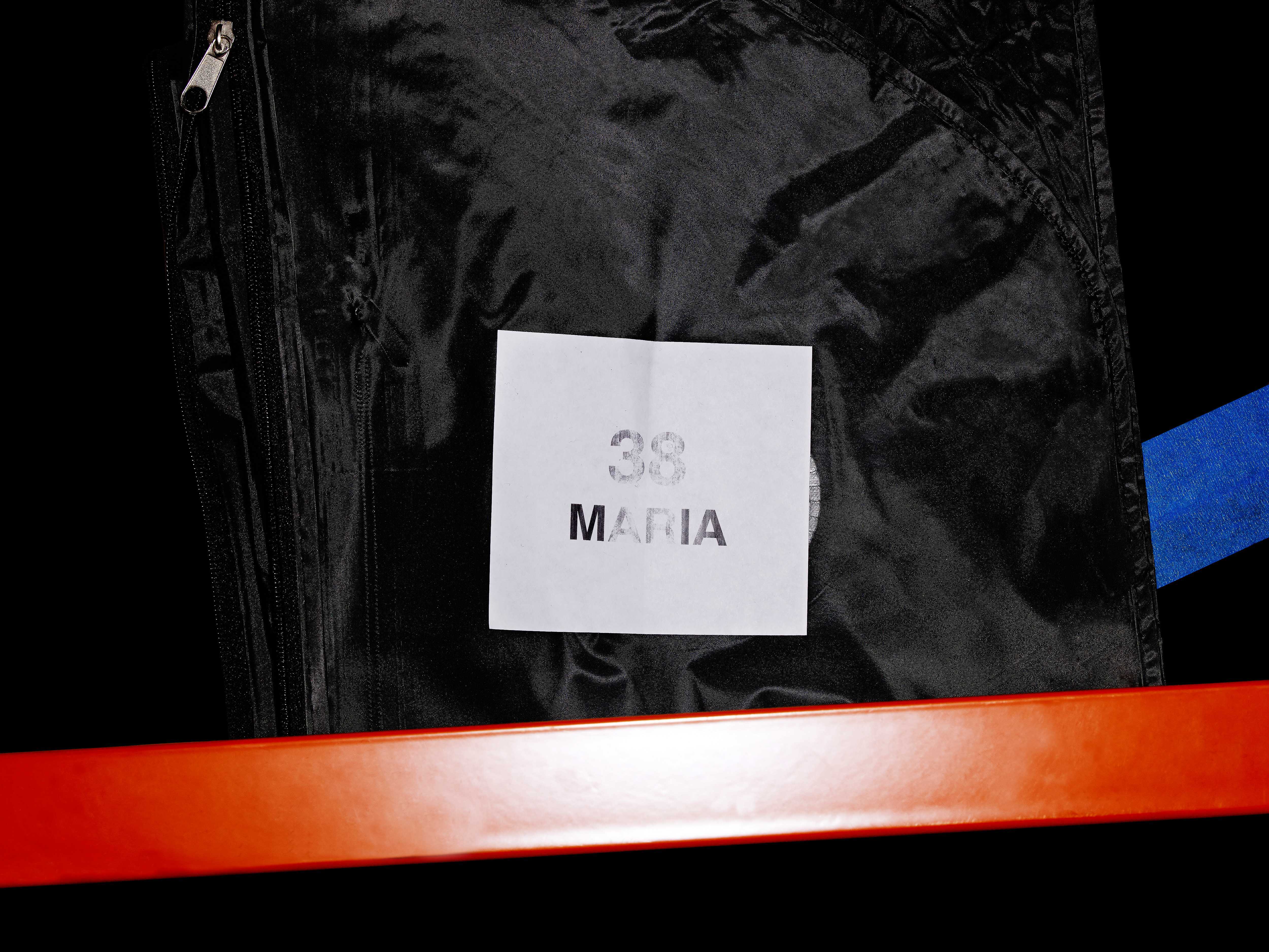 Close-up of a black garment bag with a white label that reads "38 MARIA", against a black background with a bright orange bar.