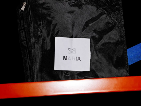 Close-up of a black garment bag with a white label that reads "38 MARIA", against a black background with a bright orange bar.
