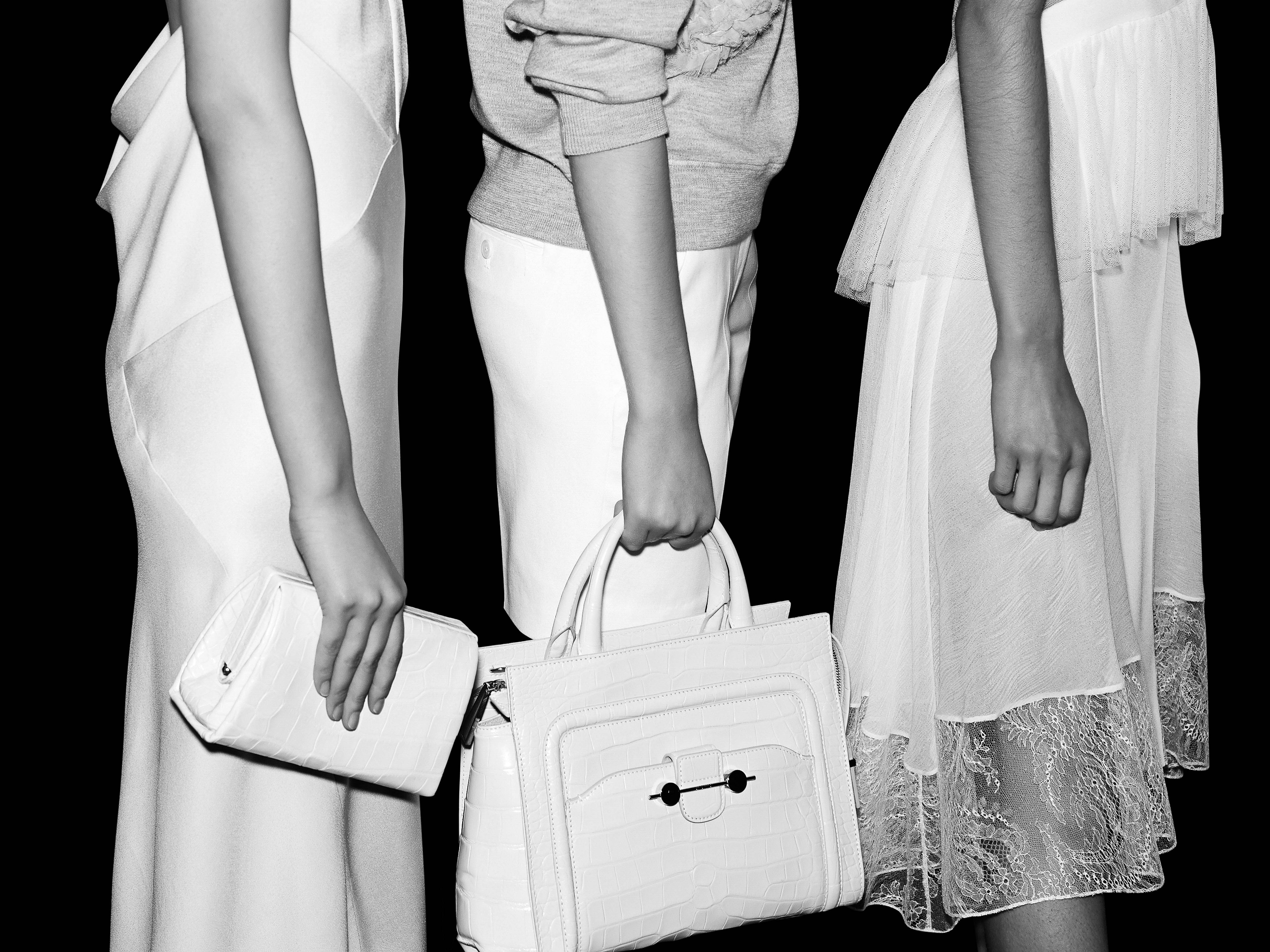 Black and white studio shot of three women in dresses holding white purses, emphasizing fashion and design elements against a dark backdrop.