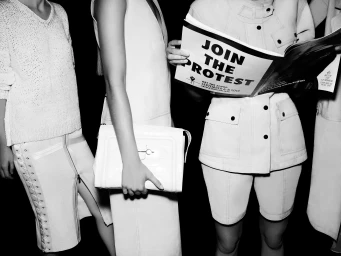 Black and white backstage shot of models in white tailored garments, with one holding a magazine reading Join The Protest.