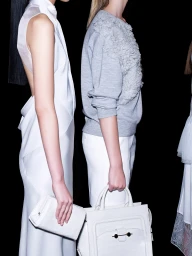 Models backstage wearing a draped white dress and grey textured sweatshirt while holding structured white leather handbags.