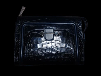 Close-up product shot of a dark embossed leather handbag with silver hardware against a black background.