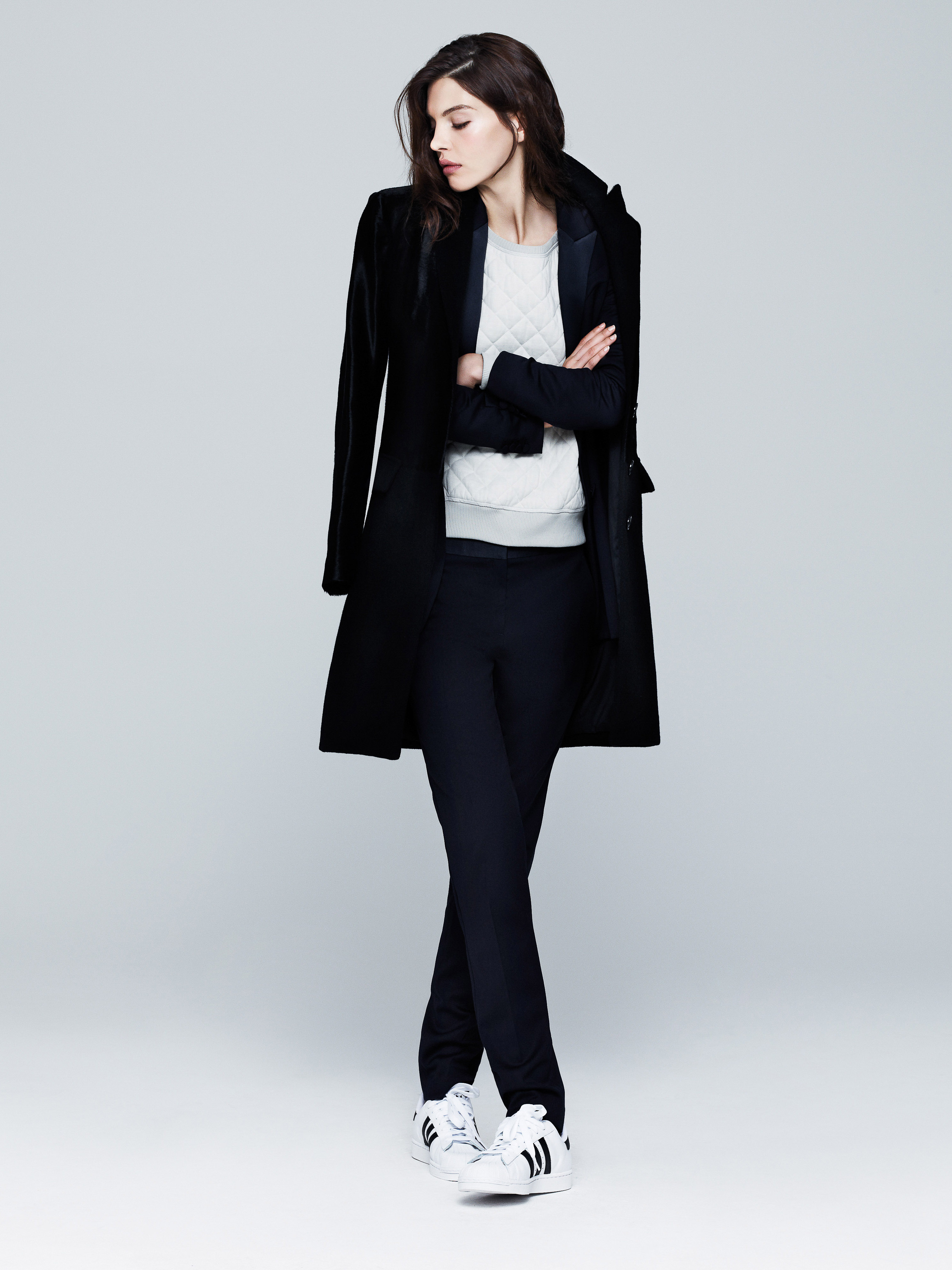 A stylish woman in a black coat and trousers stands with arms crossed, showcasing a blend of athletic and formal fashion.