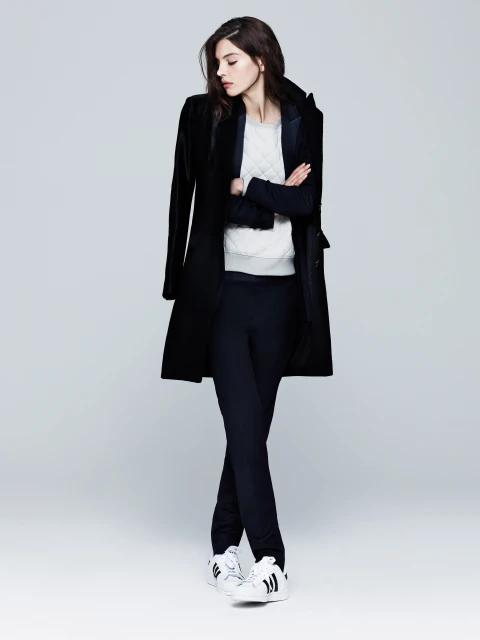 A stylish woman in a black coat and trousers stands with arms crossed, showcasing a blend of athletic and formal fashion.