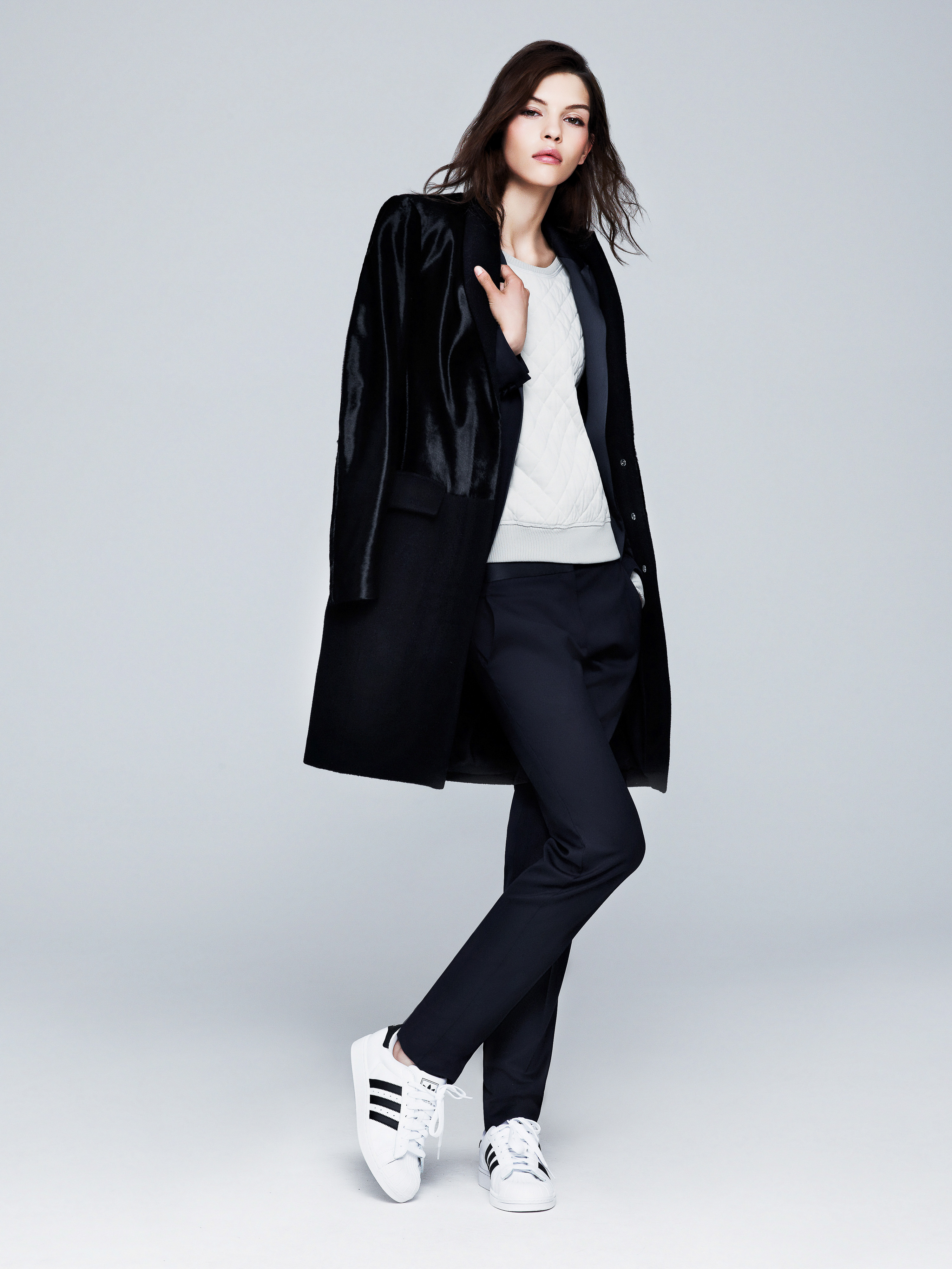 Model wearing a black tailored coat draped over a light sweater, dark trousers, and white sneakers against a grey studio backdrop.
