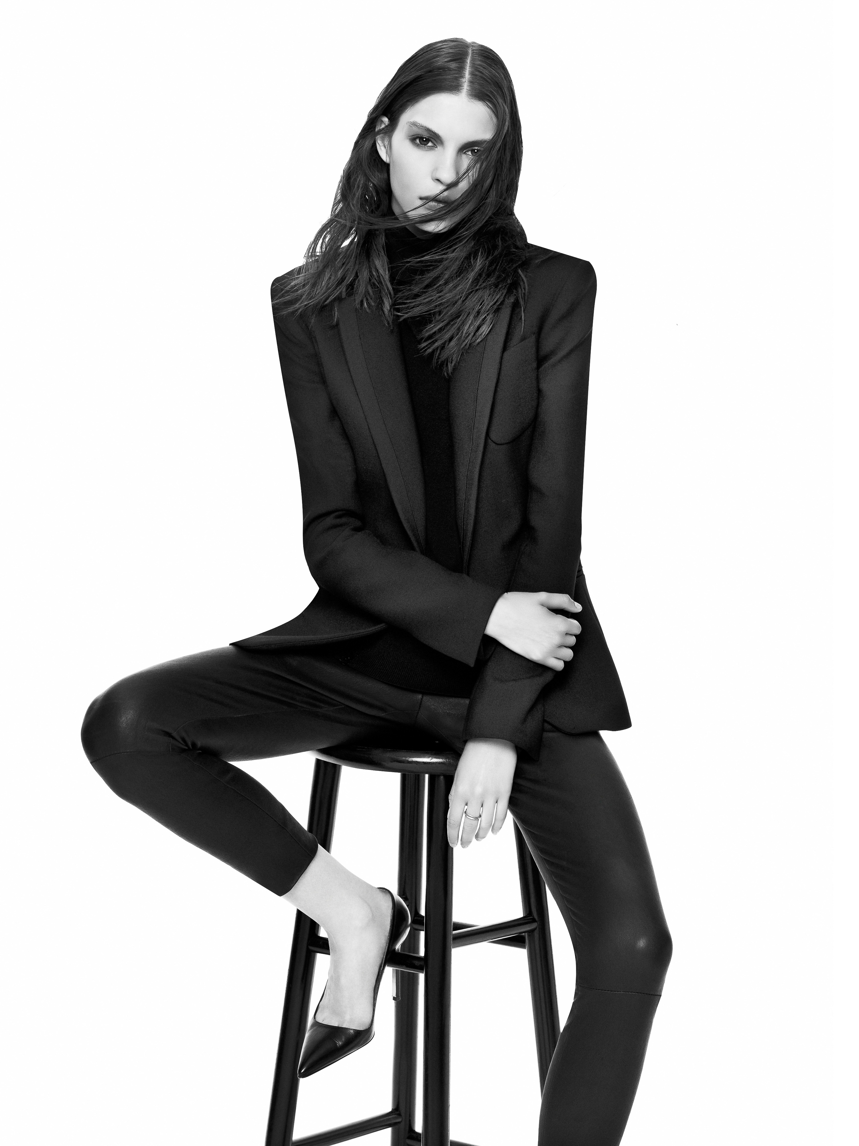 A woman in a black suit and black heels sits on a black stool against a stark white backdrop in a studio setting, showcasing fashion and style.