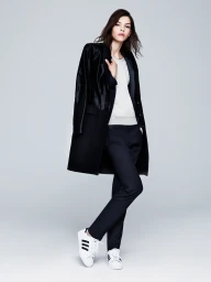 Model wearing a black tailored coat draped over a light sweater, dark trousers, and white sneakers against a grey studio backdrop.