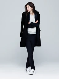 Model wearing a black tailored coat, quilted grey top, black trousers, and white sneakers against a neutral grey studio background.