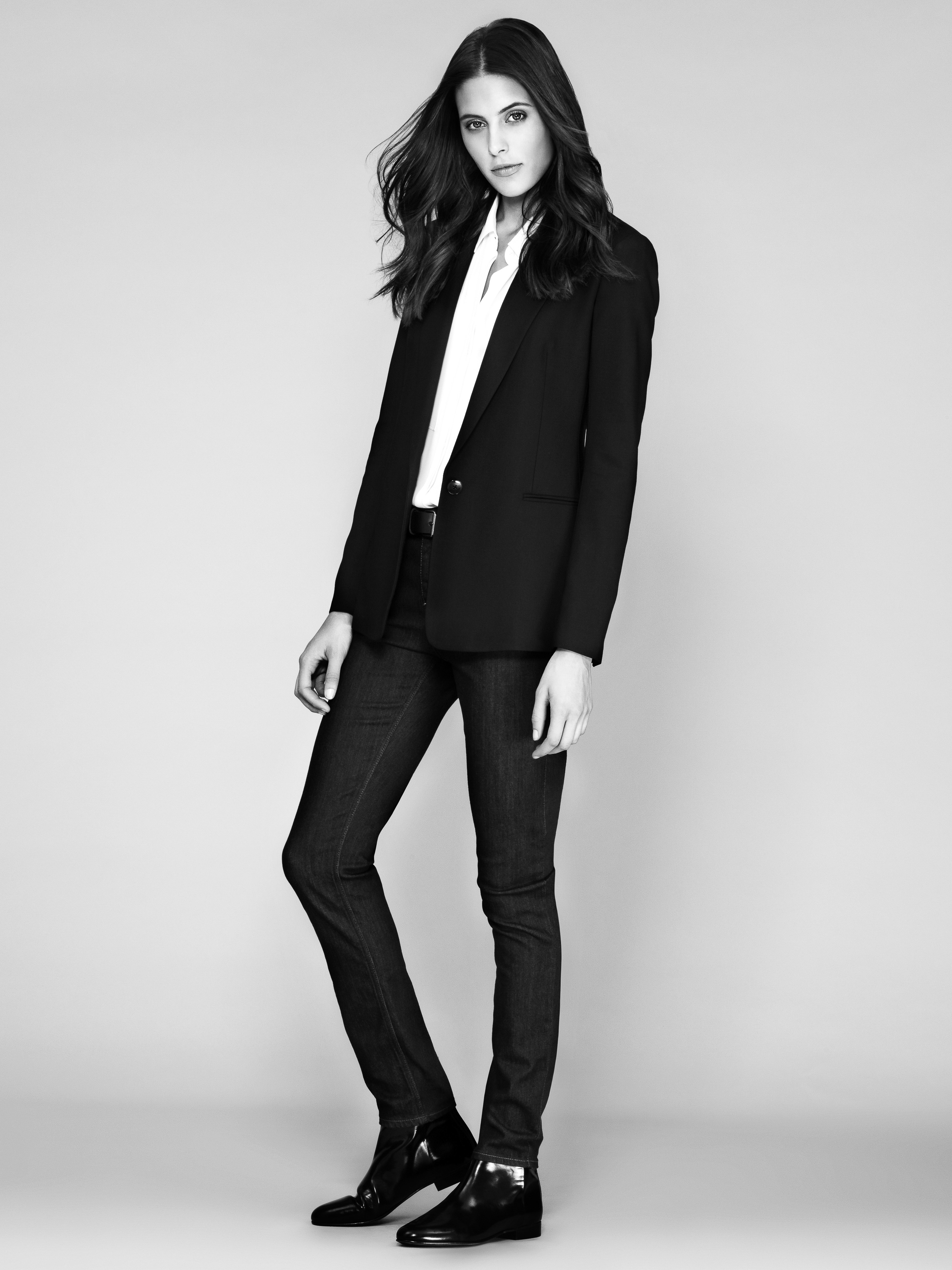 Full-length shot of a young woman in a black blazer, white shirt, and dark denim jeans against a light grey studio backdrop, fashion.