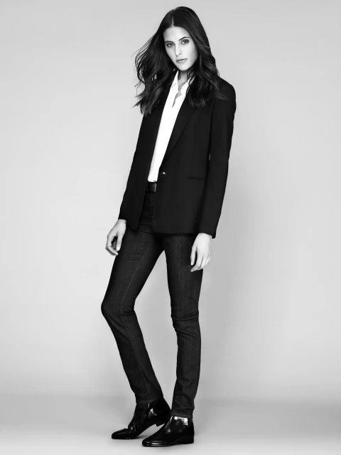 Full-length shot of a young woman in a black blazer, white shirt, and dark denim jeans against a light grey studio backdrop, fashion.