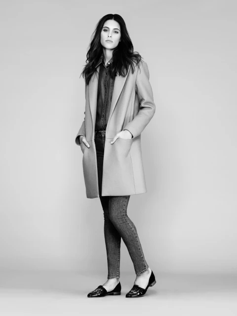 Full-length black and white portrait of a woman posing in a coat, denim shirt, jeans, and loafers against a grey background.