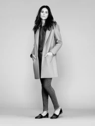 Model wearing an open mid-length tailored coat, button-up shirt, skinny trousers, and pointed loafers against a studio backdrop.