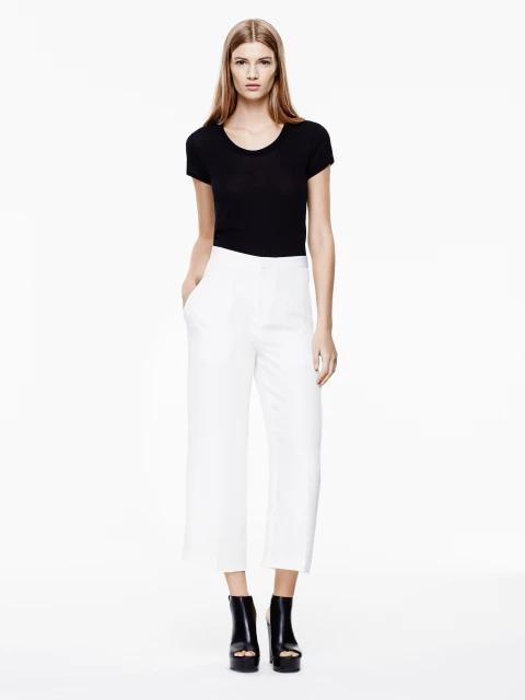 Full-length studio shot of a woman in a black top and white pants, hands in pockets, against a white backdrop, fashion photography.