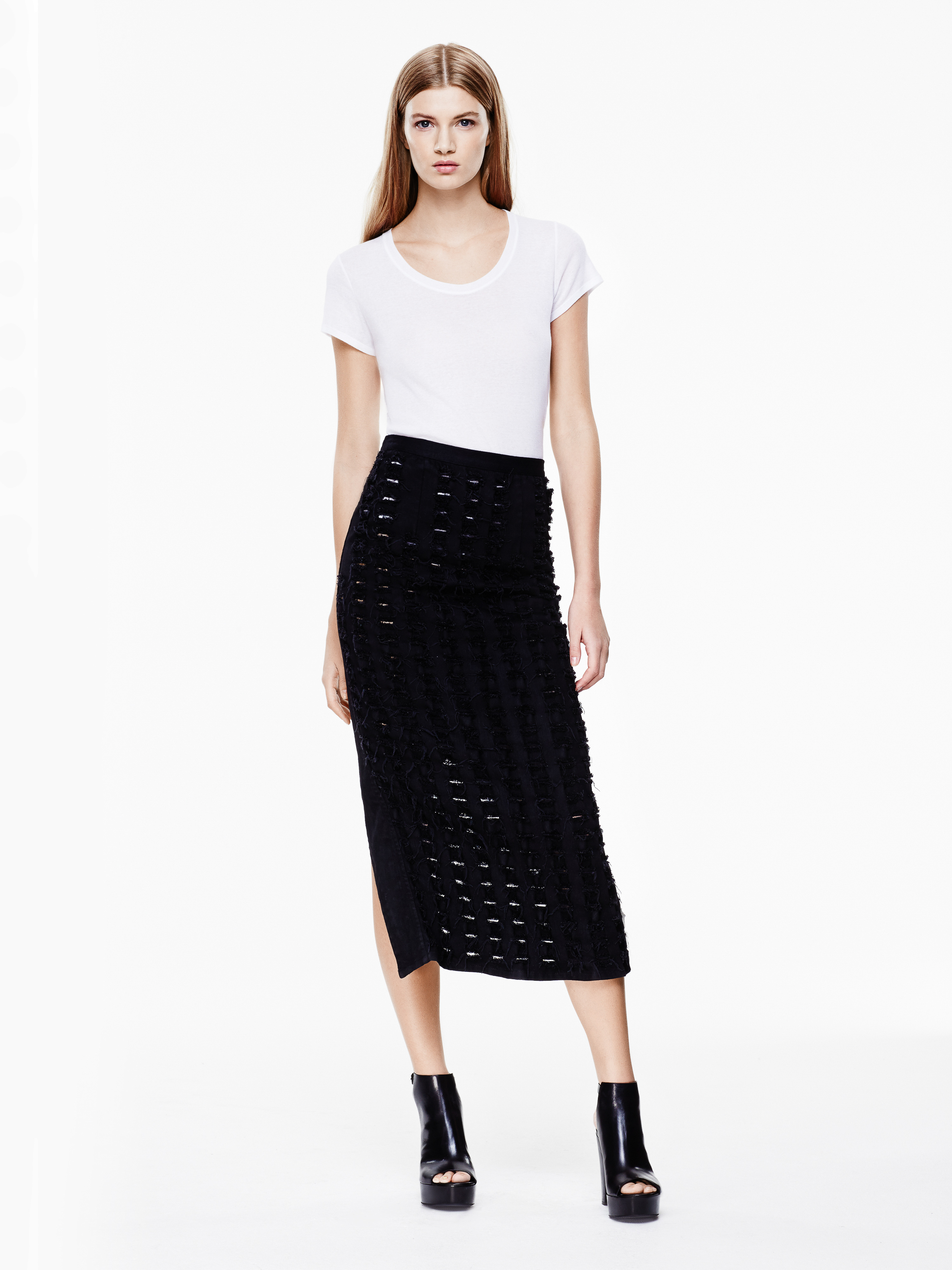 Fashion model poses in a white t-shirt and textured black midi skirt with platform heels against a white background in a studio shot.