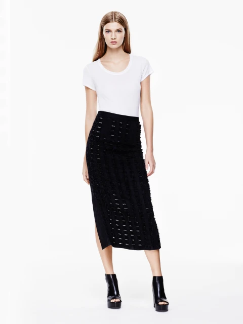 Fashion model poses in a white t-shirt and textured black midi skirt with platform heels against a white background in a studio shot.