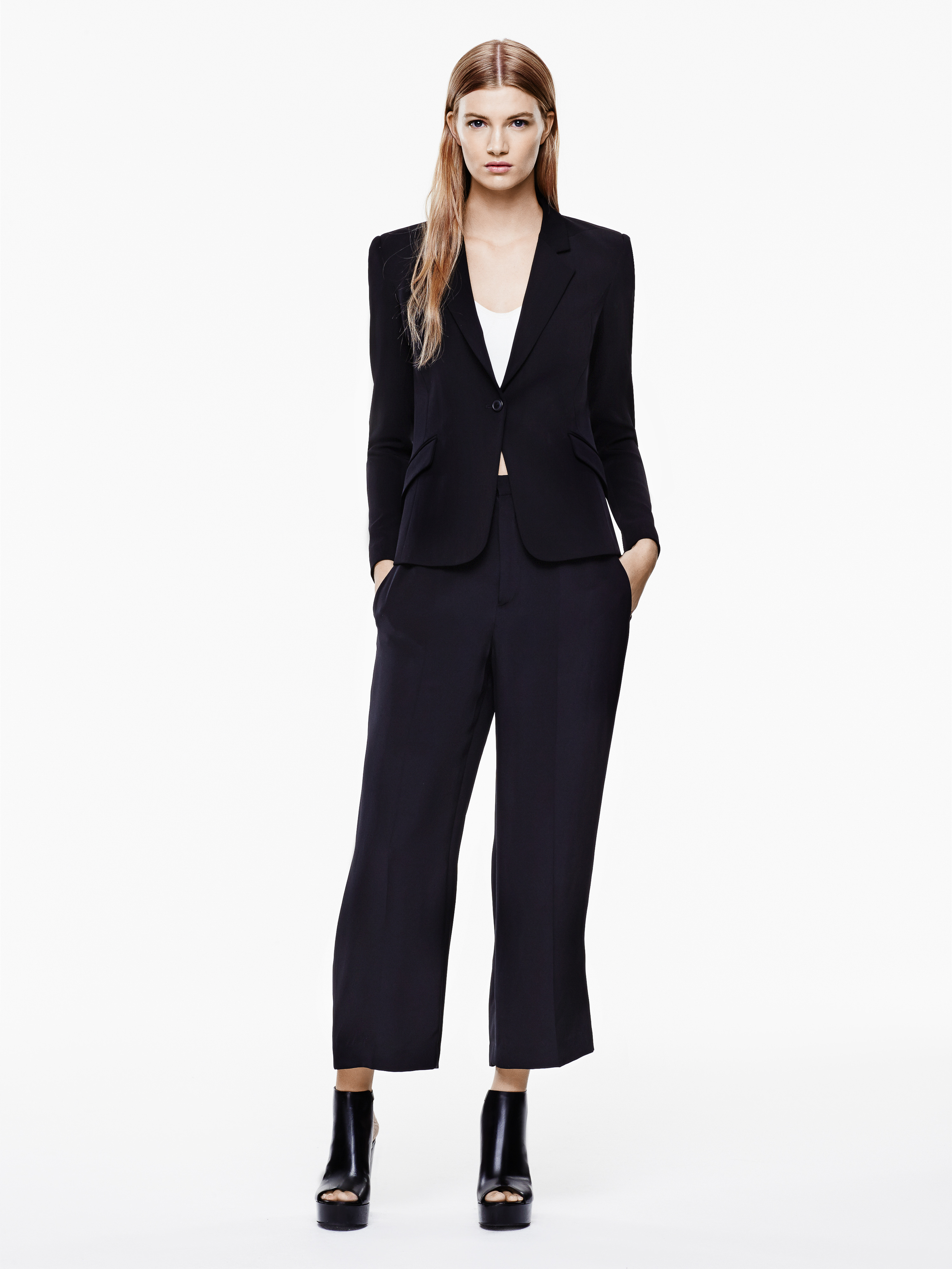 Full-length studio shot of a blonde female model in a black blazer, wide-leg pants, and platform heels on a white backdrop, fashion.