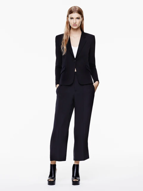 Full-length studio shot of a blonde female model in a black blazer, wide-leg pants, and platform heels on a white backdrop, fashion.