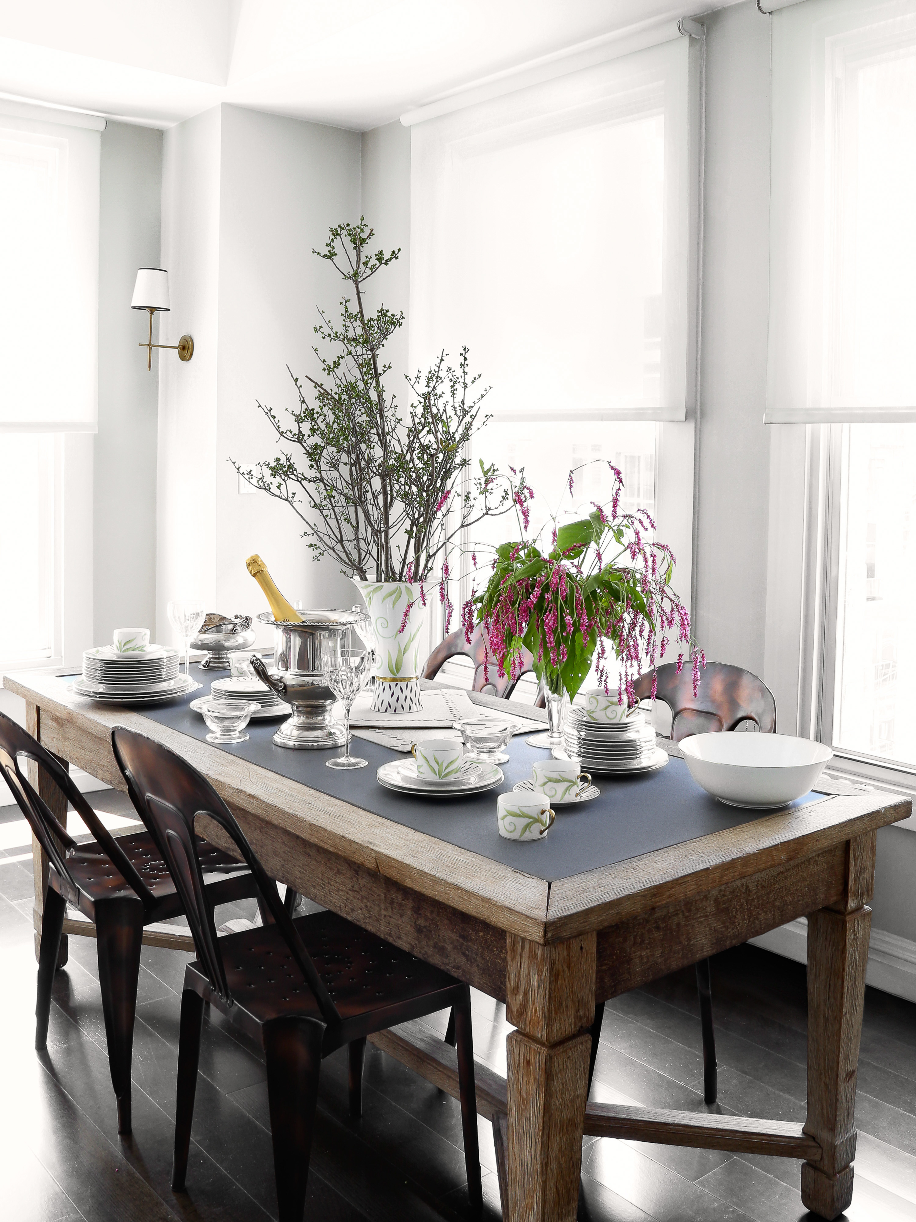Dining room table setting with champagne, flowers, dishes, glasses, and chairs under natural light coming through window blinds.
