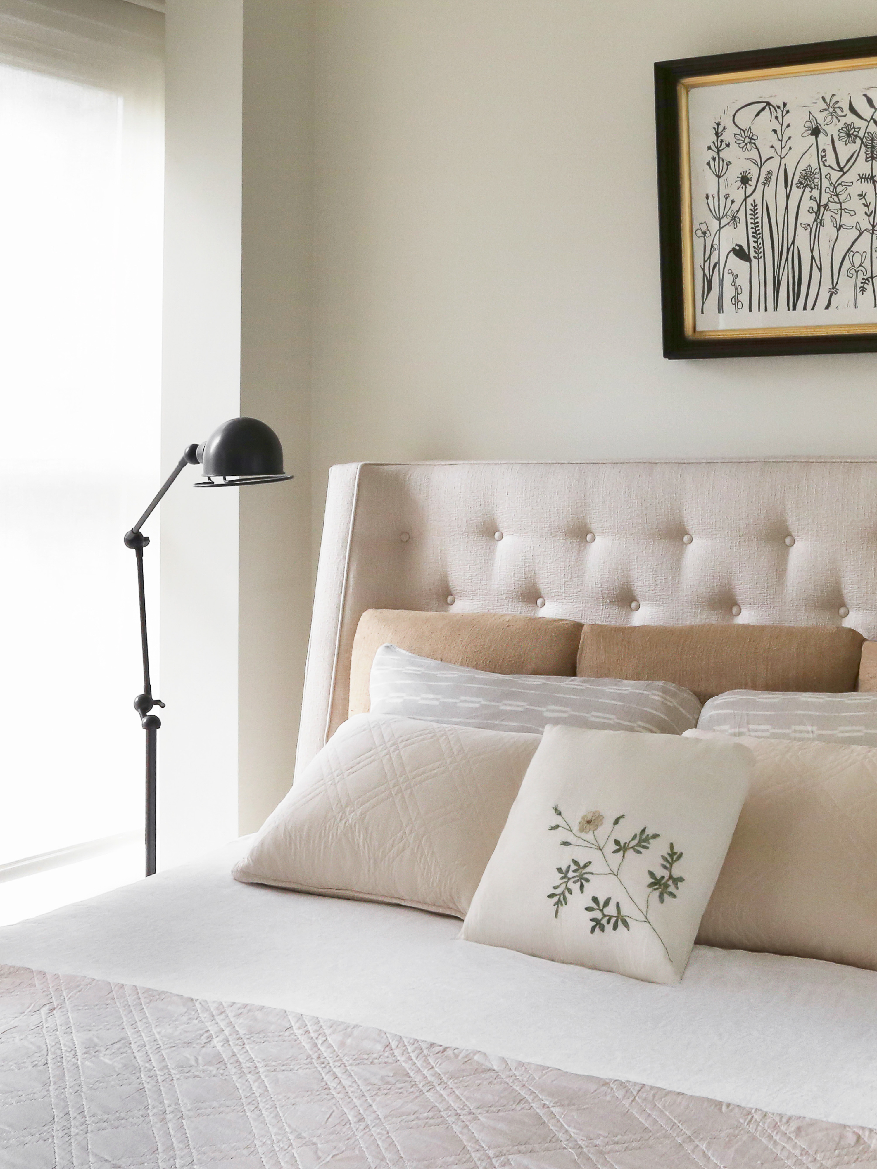 Bedroom interior featuring a neutral color palette, a tufted headboard, multiple pillows, and a black adjustable lamp.