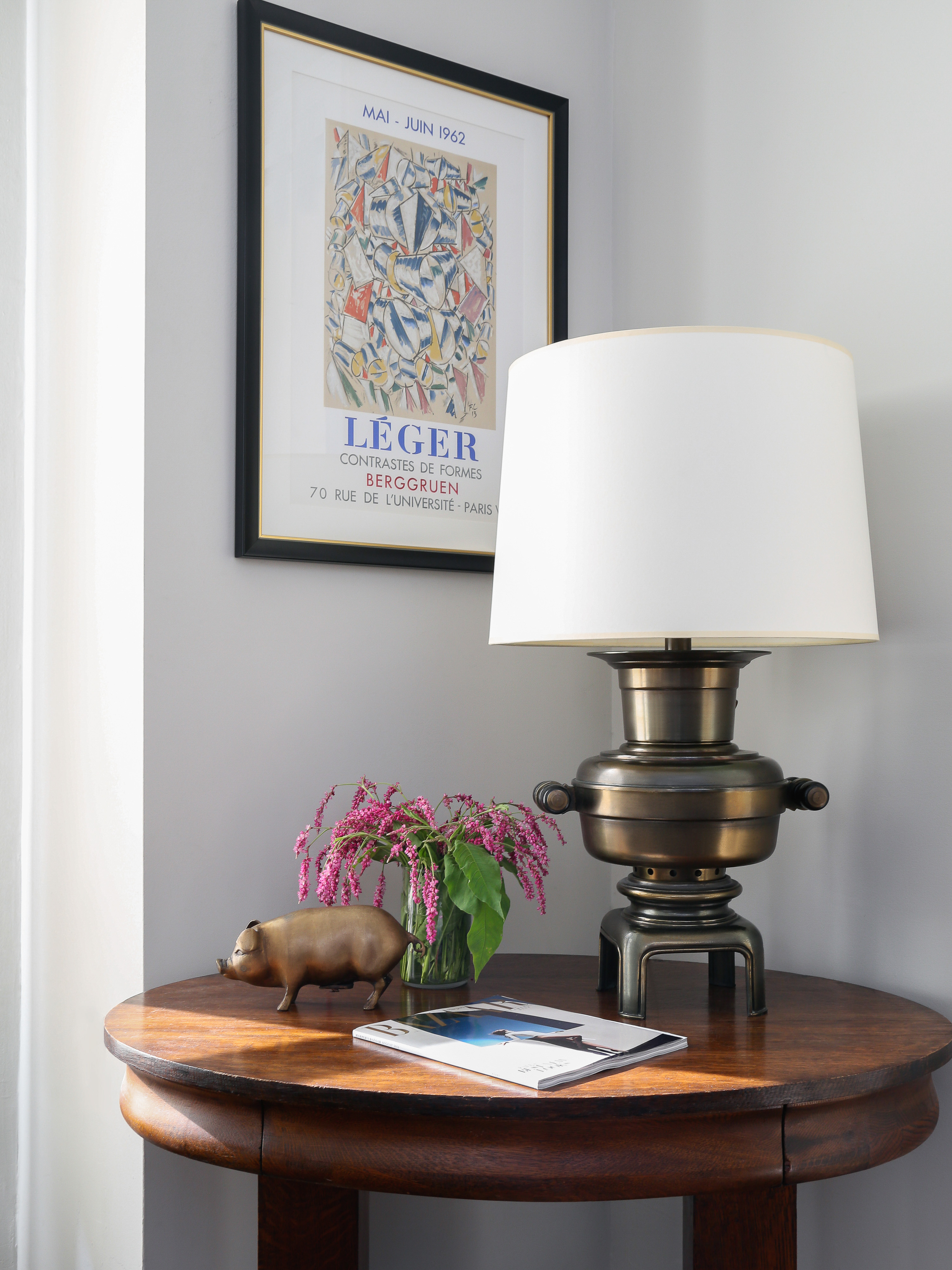 Interior view of a round wooden table with a lamp, flowers, a pig figurine, and a magazine next to a framed art print on the wall.