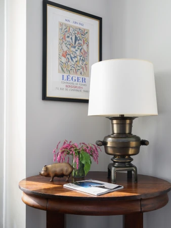Interior view of a round wooden table with a lamp, flowers, a pig figurine, and a magazine next to a framed art print on the wall.