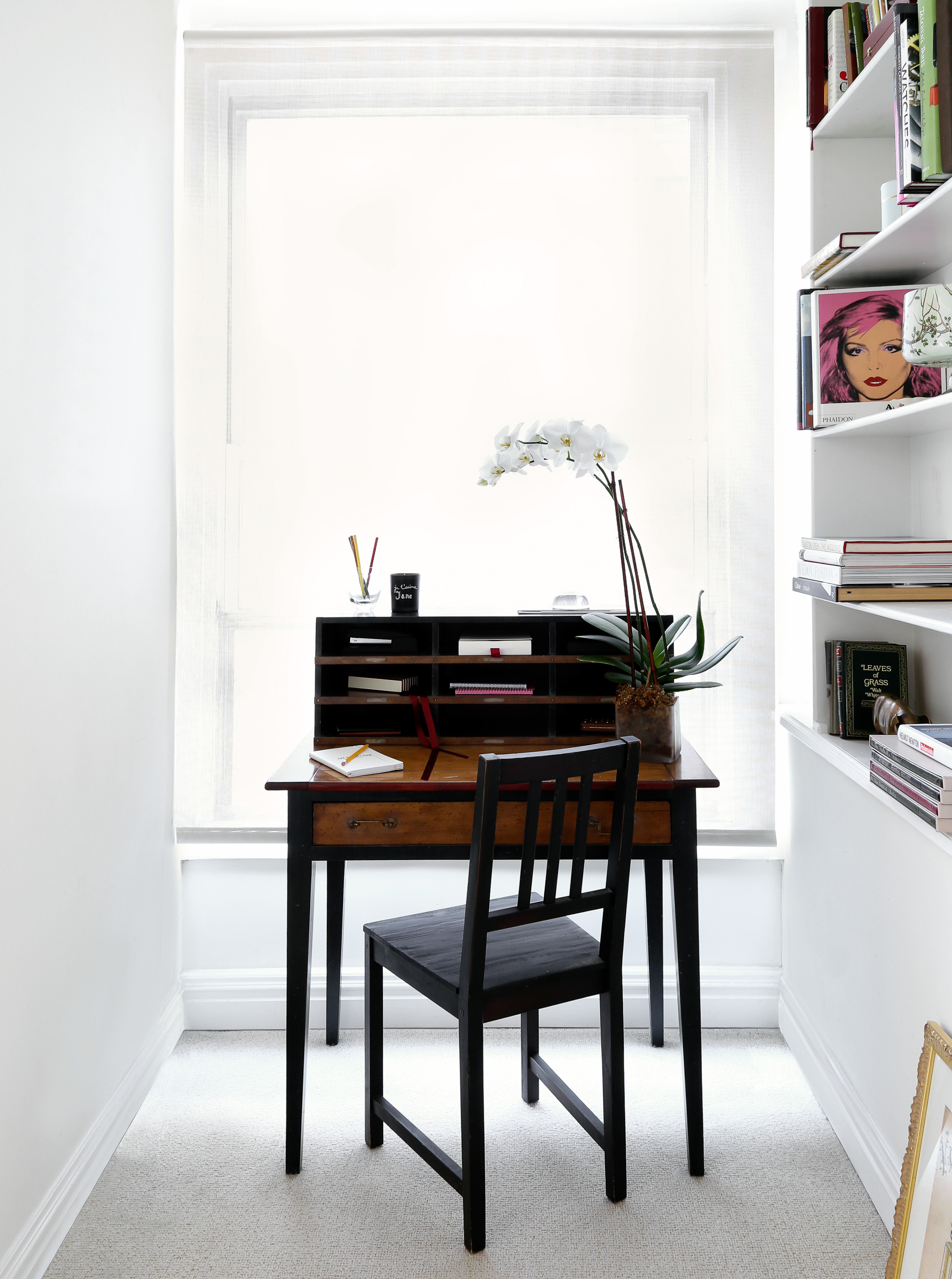 A home office area features a wooden desk and chair set against a bright window and a white wall with a bookshelf. 