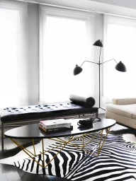Black oval coffee table with a gold wire base and a black leather daybed set over a zebra print rug in a brightly lit room.
