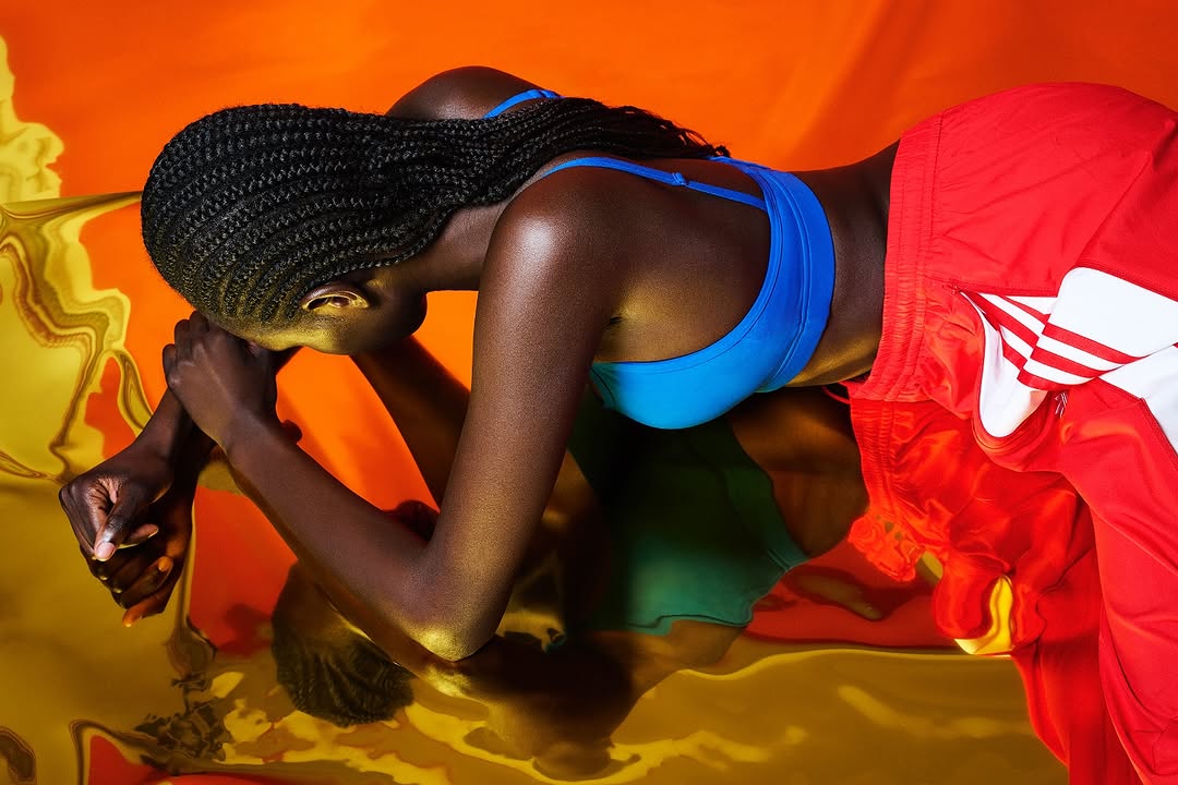 Athletic Black woman in blue sports bra and red track pants leans on reflective surface with warm orange background.