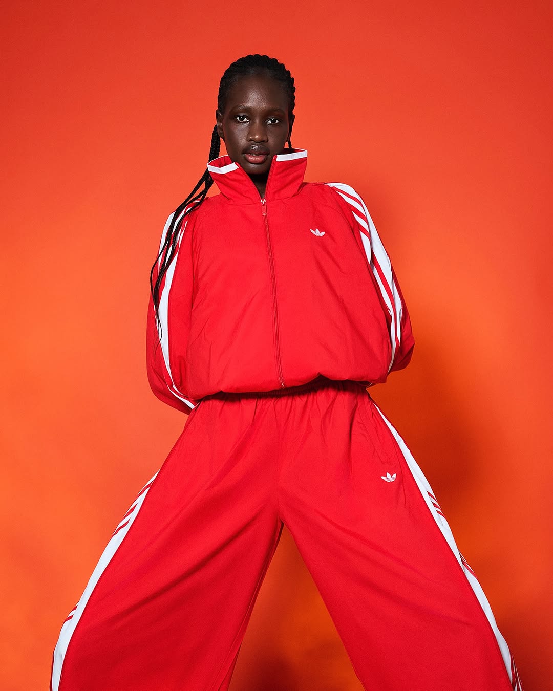 An adult fashion model poses in a red Adidas tracksuit with white stripes on a bright orange backdrop in a studio shot.