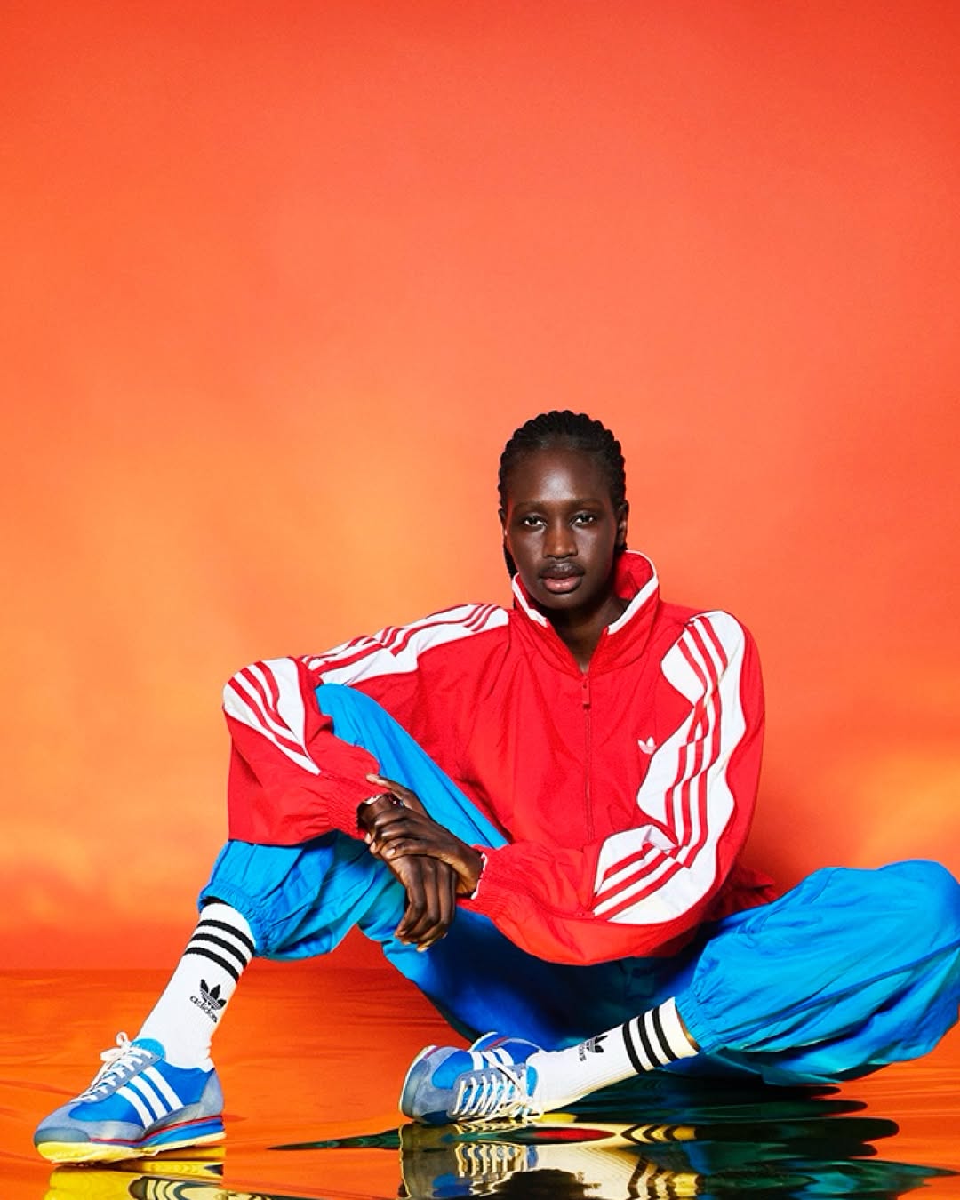 Person seated wearing a red Adidas tracksuit jacket, blue track pants, striped socks, and Adidas sneakers against an orange backdrop.