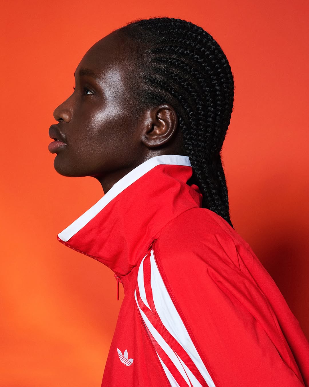 Portrait of a Black woman in profile wearing a red Adidas track jacket against an orange backdrop in a studio environment.