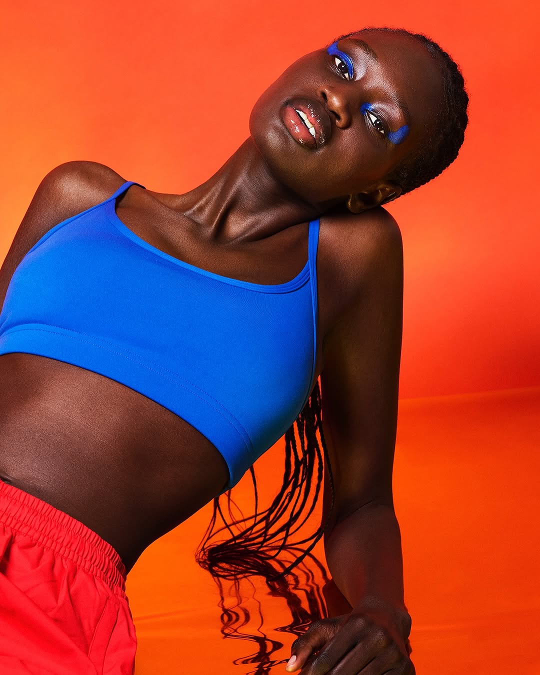 An adult Black woman is posing in a blue crop top and red shorts against an orange background with blue eyeshadow.