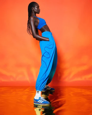Woman in blue athletic wear poses on orange reflective surface in a studio lit setting. Adidas logo is visible on socks and shoes.