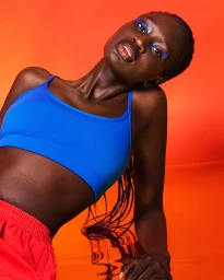 Black model with blue eyeshadow wearing a blue sports bra against vibrant orange background