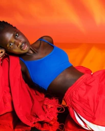 Model in a bright blue sports bra and red tracksuit against an intense orange and yellow gradient background.